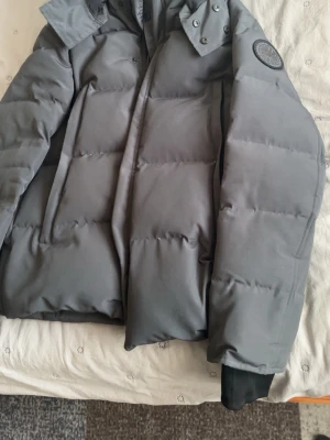 Grå jacka från Canada Goose - Snygg grå dunjacka från Canada Goose med hög krage, avtagbar huva och diskret logotyp på ärmen. Jackan har breda kanaler, dolda fickor och muddar vid ärmsluten för extra värme. Perfekt för kalla vinterdagar och riktigt stilren.