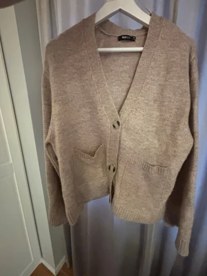 Beige kofta från Gina Tricot  - Mysig beige kofta från Gina Tricot med v-ringning, stora knappar framtill och två praktiska fickor. Koftan är stickad i mjukt material och har en avslappnad passform med ribbade muddar vid ärmslut och nederkant. Perfekt för lager på lager och enkel att styla. Storlek M men passar mig som brukar bära Xs/S