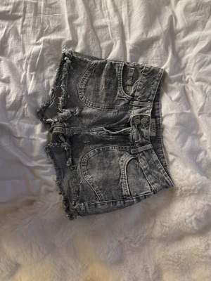 Grå jeansshorts med fransar från Lindex - Säljer ett par grå jeansshorts från Lindex med slitna detaljer och fransig kant. Shortsen har hög midja, klassiska fem fickor och dubbla silverknappar i midjan. Perfekta för en avslappnad och trendig look.