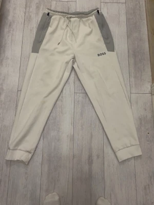 Beige mjukisbyxor från BOSS - Snygga beige mjukisbyxor från BOSS med grå detaljer vid fickorna och midjan. Byxorna har resår i midjan med dragsko och muddar vid bensluten. Tillverkade i mjukt material för maximal komfort. Perfekta för en avslappnad stil.