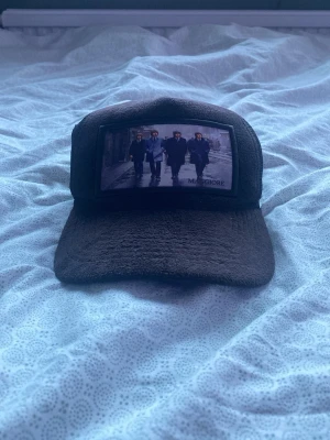 Svart truckerkeps Maggiore - Säljer en svart truckerkeps från Maggiore med mesh på sidorna och ett stort tryck framtill som visar fyra personer i kostym. Kepsen har böjd skärm och ett broderat M-logotyp på sidan. Perfekt för dig som vill sticka ut med en unik accessoar.