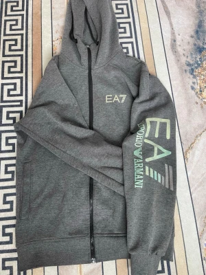 Grå EA7 Emporio Armani hoodie - Snygg grå hoodie från EA7 Emporio Armani med dragkedja framtill och huva. Stora logotryck på bröstet och ärmen i vitt och guldigt som har tappt färg och blivit  ljusgrönt. Den sitter jätte bra och luvan sitter jätte fint. hör gärna av er om ni har några frågor elr funderingar 