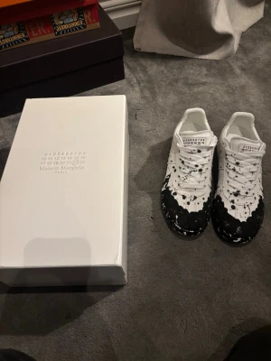 Maison Margiela svartvita sneakers - Maison Margiela sneakers i vitt skinn med svart färgstänksmönster. Klassisk rund tå, snörning och platt sula. Kommer med originalkartong och skyddspåsar. Unik design som sticker ut och passar perfekt till streetwear. Passar 40 men storlek 39. Kom dm om ni har ni har frågor 