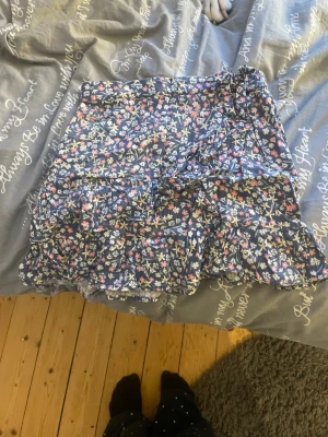 Blommig volangkjol med knyt - Söt kortkjol med blommigt mönster i blått, rosa, vitt och gult. Kjolen har volangdetaljer och ett Rosetband i midjan som ger en extra touch. Perfekt för dig eller din dotter som gillar en romantisk och trendig stil.