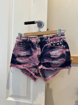 Rosa och blåa slitna jeansshorts - Coola jeansshorts i rosa och blå med slitna detaljer och nitar på fickan. Shortsens råa kanter och tuffa tvätt ger en edgy vibe. Perfekta för dig som vill sticka ut med din stil i sommar.