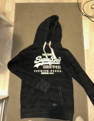Mörkblå Superdry hoodie med tryck - Snygg mörkblå hoodie från Superdry med vitt tryck på bröstet och japanska tecken. Hoodien har vita dragsnören och en klassisk känguruficka framtill. Perfekt för en avslappnad streetwear-look.