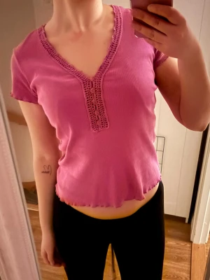 Rosa croppad topp med spetsdetaljer - Söt rosa croppad topp med v-ringning och spetsdetaljer längs halsringningen. Toppen har korta ärmar och vågig kant nertill, perfekt för en trendig och avslappnad look.