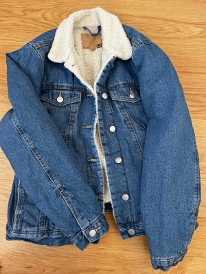 Blå fodrad jeansjacka från 157 Stl S - Snygg blå jeansjacka från 157 med vit teddyfoder på insidan och kragen. Klassisk modell med två bröstfickor med lock och silverfärgade knappar framtill. Perfekt för dig som vill ha en cool och varm jacka med streetkänsla.