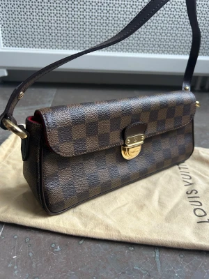 Louis Vuitton axelremsväska modell Ravello vintage - Snygg axelremsväska från Louis Vuitton i klassiskt brunt Damier-mönster. I princip oanvänd. Väskan har guldfärgat lås framtill, röd insida och axelrem i mörkbrunt läder. Tillverkad i canvas med läderdetaljer och kommer med original dustbag. Authentic stämpel inuti. Köpt av mig i LV butik. Mått: längd 27 cm, höjd 12 cm, bredd 7 cm