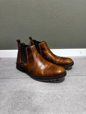Rieker Chelsea boots i brunt läder  - Ett par bruna Chelsea boots med resårpaneler på sidorna. Skorna har en robust sula och en dragflik baktill för enklare påtagning. De är designade för att vara lätta att matcha med olika outfits.