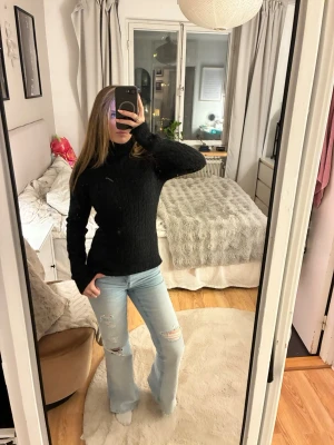 Svart kabelstickad polotröja - En svart kabelstickad polotröja med lång ärm och hög krage. Tröjan har en figurnära passform och klassiskt mönster. Perfekt för att hålla sig varm under kyliga dagar. Materialet ser ut att vara mjukt och bekvämt.