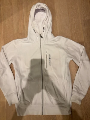 Vit hoodie från Sail Racing - Säljer en stilren vit hoodie från Sail Racing med dragkedja framtill och en extra ficka med dragkedja på bröstet. Hoodien har justerbar huva med snören och logga broderad på bröstet. Perfekt för dig som gillar sportig och clean stil. Skriv för frågor 💗