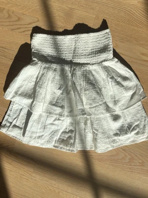 Vit volangkjol från Kappahl - Söt vit kjol från Kappahl med bred smock i midjan och dubbla volanger. Kjolen är gjord i ett lätt, luftigt tyg som ger en avslappnad och somrig känsla. Perfekt att matcha med en enkel topp för en fräsch look.