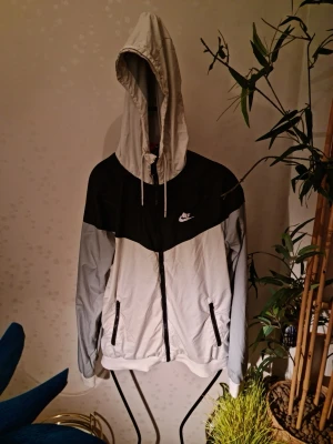 Nike svartvit vindjacka med huva - Nike vindjacka i svart och vitt med dragkedja, huva och två fickor framtill. Jackan har en sportig look med logga på bröstet och justerbara snören vid huvan. Perfekt för blåsiga dagar och streetwear-stil.