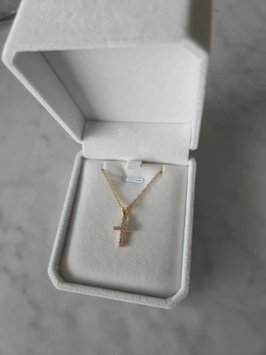 Guldfärgat halsband med korshänge - Snyggt halsband i guldfärg med ett korsformat hänge dekorerat med små glittrande pärlor. Kedjan är tunn och elegant, perfekt för att ge en stilren touch till din outfit. 