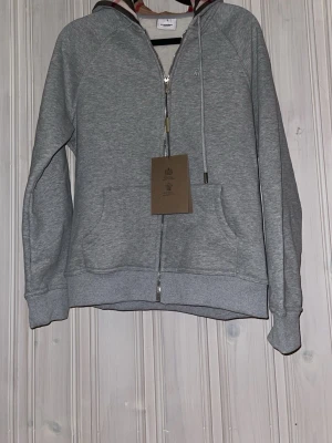 Grå hoodie med dragkedja från Burberry - Snygg grå hoodie från Burberry med dragkedja framtill och klassiskt rutigt foder i huvan. Hoodien har två stora fickor fram, ribbade muddar och justerbar dragsko i huvan. Perfekt för en chill och stilren look.