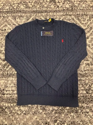Mörkblå kabelstickad tröja Polo Ralph Lauren - Mörkblå kabelstickad tröja från Polo Ralph Lauren med rund halsringning och röd broderad logga på bröstet. Tröjan har långa ärmar och ribbstickade muddar vid ärmslut och nederkant. Perfekt för en stilren och klassisk look. Tröjan är i storlek S men passar även för folk i storlek M. Hör av dig vid eventuellt frågor eller funderingar! 