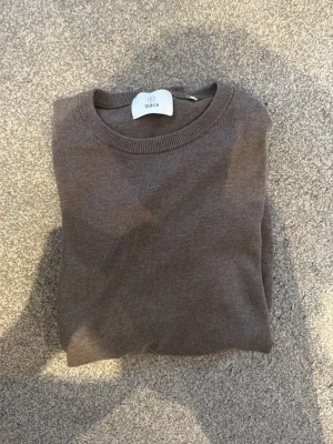 Brun stickad tröja från Bläck - Mjuk och stilren brun stickad tröja från Bläck med rund halsringning och ribbade muddar. Perfekt för lager-på-lager och enkel att matcha med jeans eller chinos.
