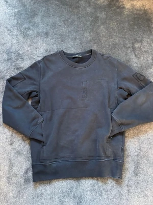 Svart sweatshirt från Stone Island - Säljer en svart sweatshirt från Stone Island med klassisk rund hals och lång ärm. Tröjan har en bröstficka och den ikoniska Stone Island-loggan på vänster ärm. Tillverkad i mjuk bomull för skön komfort och stilren look. Ser lite dammig ut men de löser jag såklart innan jag skickar den. 