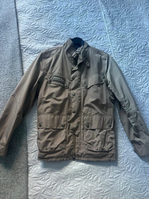 Beige vindjacka från Barbour - Snygg beige vindjacka från Barbour International med patch på bröstet, fyra framfickor med tryckknappar och hög krage med spänne. Jackan har en klassisk rak passform och är tillverkad i ett lätt, vindtätt material som passar perfekt till streetwear.