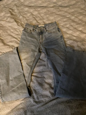 Ljusblå utsvängda jeans med låg midja - Säljer ett par ljusblå jeans med bootcut passform och låg midja. Jeansen har klassisk femficksdesign och gylf med dragkedja. Perfekta för en avslappnad och trendig look. Materialet är denim i bomull.