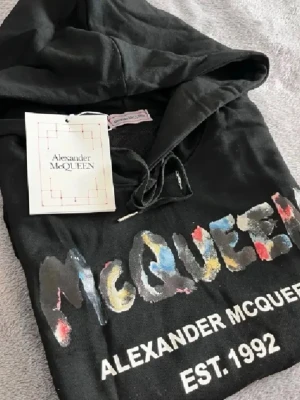 Svart hoodie Alexander McQueen - Svart hoodie från Alexander McQueen med färgglad logga framtill och texten 'EST. 1992'. Hoodien har huva med dragsko och en stor magficka. Materialet ser ut att vara mjukt bomullstyg, perfekt för en avslappnad streetwear-look.