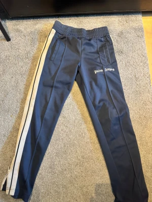 Palm Angels byxor  - Säljer ett par blåa mjukisbyxor från Palm Angels med vita sidoränder och logga på vänster ben. Byxorna har resår i midjan och raka ben. Perfekta för en avslappnad och trendig look. Äkata såklart!
