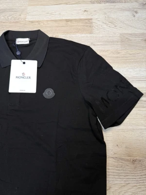 Svart Moncler pikétröja - Säljer en svart pikétröja från Moncler med klassisk krage och korta ärmar. Tröjan har Moncler-logga på bröstet och diskret Moncler-tryck på ärmen. Tillverkad i mjuk bomull för en bekväm känsla och stilren look.