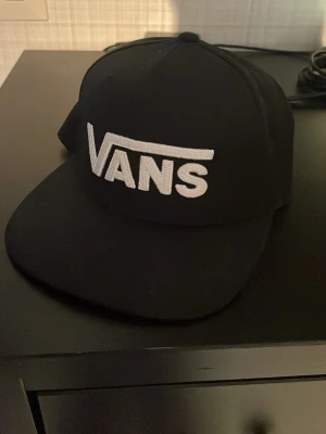 Svart keps från Vans med broderad logga - Säljer en svart keps från Vans med stor vit broderad logga framtill. Klassisk snapback-modell med justerbar plastrem bak. Perfekt för dig som gillar streetwear och vill ha en stilren accessoar.