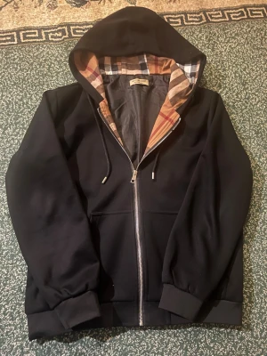 Svart hoodie från Burberry - Snygg svart hoodie från Burberry med dragkedja och klassiskt rutigt foder i huvan. Hoodien har ribbade muddar, fickor framtill och justerbar dragsko i huvan. Perfekt för dig som vill ha en stilren och exklusiv look med ikoniska Burberry-detaljer.