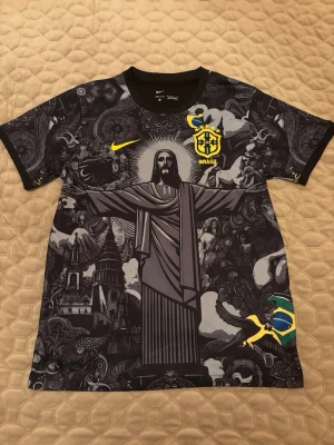 Svart Brasil Nike topp med tryck - Unik svart topp från Nike med Brasil-logga och stort grafiskt tryck av Kristusstatyn och brasilianska flaggan. T-shirten har rund hals, korta ärmar och detaljer i gult. Perfekt för dig som vill sticka ut med en sportig vibe.