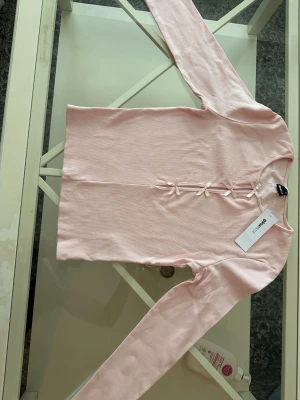 Ljusrosa ribbad topp med rosetter - Supersöt ljusrosa långärmad topp från Gina Tricot med ribbad struktur och dekorativa små rosetter framtill. Rund halsringning och mjukt stretchigt material som sitter bekvämt. Perfekt för en gullig och trendig look.