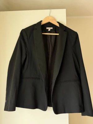 Svart klassisk kavaj från H&M - Storlek 46 men passar mig som har STORLEK S. Stilren svart kavaj från H&M med klassisk passform och två fickor framtill. Kavajen har smala slag och enkelknäppning, perfekt för en clean och modern look. Tillverkad i ett mjukt tyg med lätt stretch för extra komfort.