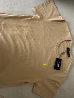 Beige t-shirt från Polo Ralph Lauren - Snygg beige t-shirt från Polo Ralph Lauren med klassisk gul broderad logga på bröstet. T-shirten har rund halsringning och korta ärmar. Tillverkad i mjuk bomull för en skön känsla och enkel stil.
