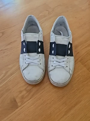 Valentino open Navy - Snygga vita sneakers från Valentino med bred svart rem över vristen och vita nitar på sidorna. Skorna har klassisk låg profil, vita skosnören och coola detaljer runt hälen. Tillverkade i läder för en exklusiv känsla och stilren look.