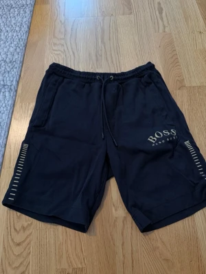Svarta shorts från Hugo Boss - Snygga svarta shorts från Hugo Boss med dragsko i midjan och logga tryckt på vänster ben. Shortsen har elastisk midja med Hugo Boss-detaljer och diskreta ränder på benen. Perfekta för en avslappnad och stilren look.