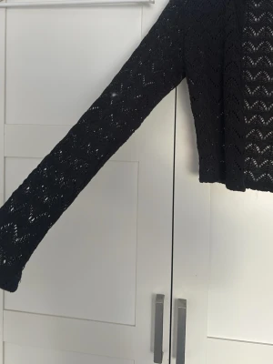Svart virkad kofta med mönster - Snygg svart kofta med virkat zigzag-mönster och dekorativa silverknappar framtill. Koftan har lång ärm och är tillverkad i ett tunt, luftigt material som ger en cool look. Perfekt att slänga över en topp för extra stil.