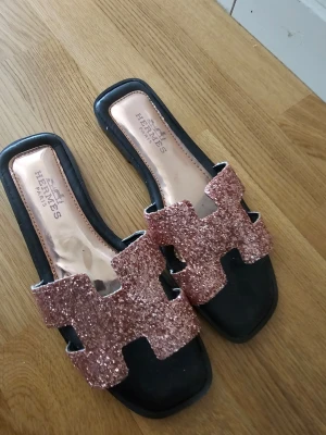 Glittriga sandaler  - Säljer ett par snygga sandaler  med breda remmar täckta av rosa glitter. Sandalerna har svart sula och öppen tå med fyrkantig form. Perfekta för dig som vill sticka ut med lite extra bling i din outfit. Använd 1 gång