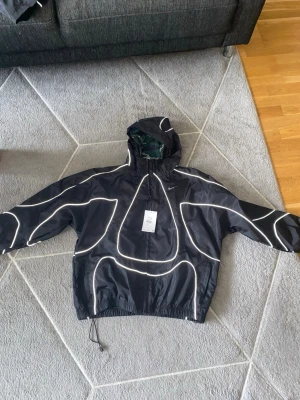 Nike svart vindjacka med reflexdetaljer - Svart vindjacka från Nike med unika vita reflexlinjer över hela jackan och huva. Jackan har dragsko i nederkant och en tryckt detalj på ryggen. Perfekt för dig som vill sticka ut och synas i mörkret. Materialet är lätt och vindtätt.