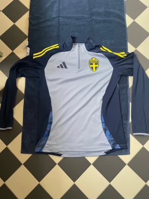 Sverige träningströja Adidas blå/gul - Snygg långärmad träningströja från Adidas i blått  med gula detaljer på axlarna. Tröjan har halv dragkedja, Sveriges landslags logga på bröstet och är tillverkad i funktionsmaterial som andas. Perfekt för träning eller uppvärmning. Använd 2-4 gånger och inget tecken på användning. 