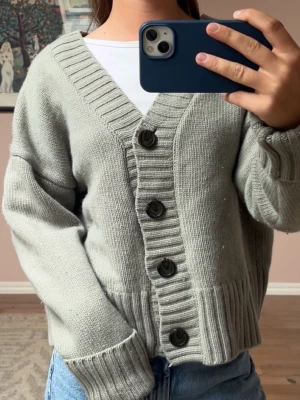 Ljusgrå stickad kofta med knappar - Mysig ljusgrön/grå stickad kofta med stora svarta knappar framtill. Koftan har bred ribbstickad kant vid ärmslut, nederkant och längs knäppningen. Oversized passform och V-ringad hals. Perfekt att slänga över en t-shirt för en avslappnad look.