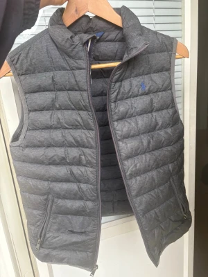 Grå väst från Polo Ralph Lauren  - Snygg grå dunväst från Polo Ralph Lauren. Säljer för att sonen växt ur den. Perfekt för sommar, vår, höst. Använt men bra/okey skick. Helhetsintrycket fint. Liten reva(se bild) därav priset . Dock tänker man inte på revan så mycket. Går att sy/tejpa. Skriv gärna om du har frågor eller vill ha fler bilder. Ungdomsstorlek L(14-16). Mått: Längd från axel till nedre kant ca 53cm. bredd där som bredast ca 43-44cm. Skickar snabbt dvs samma dag eller nästa dag. Mvh Mimmi