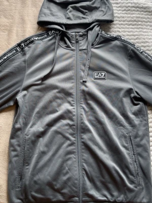 Grå EA7 Emporio Armani hoodie - Snygg grå hoodie från EA7 Emporio Armani med dragkedja, huva och dragsnören. Svarta detaljer med logga längs axlarna och EA7-märke på bröstet. Tillverkad i mjukt material som känns skönt mot huden. Perfekt för en chill och sportig look.