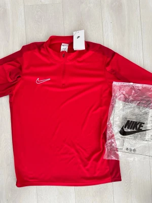 Röd träningströja från Nike - Riktigt snygg röd träningströja från Nike med halv dragkedja och vit broderad swoosh på bröstet. Tillverkad i ett lätt och funktionellt material som andas, perfekt för träning eller sportaktiviteter. Tröjan har en sportig passform och hög krage.