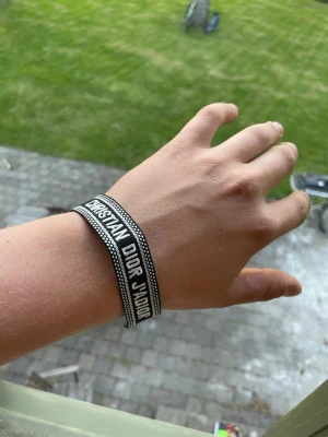 Christian Dior J'Adior armband svart - Snyggt svart tygarmband från Christian Dior med texten 'J'Adior' i vitt. Armbandet har en vävd design med vita detaljer längs kanterna och justerbar knytning. Perfekt accessoar för dig som gillar exklusiva märken och vill sticka ut.
