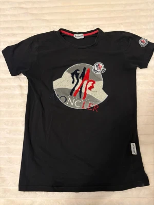 Svart Moncler t-shirt med logga - Snygg svart t-shirt från Moncler med stor broderad logga i vitt, rött och blått på bröstet samt patch på ärmen. Klassisk rund hals och korta ärmar. Perfekt för dig som gillar streetwear och vill ha något stilrent men ändå med en twist.