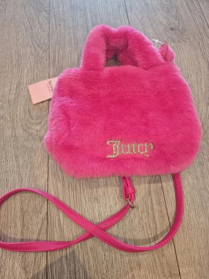 Juicy Couture rosa fluffig handväska - Supergullig rosa fluffig handväska från Juicy Couture med glittrande Juicy-logo framtill. Väskan har både handtag och en lång axelrem i matchande rosa färg. Perfekt accessoar för dig som vill sticka ut med en färgstark och mjuk look.