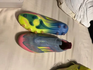 Adidas F50 blå/rosa fotbollsskor - Säljer ett par Adidas F50 fotbollsskor i blått och rosa med gula detaljer. Skorna har en strömlinjeformad design utan snörning och en sula i neonfärger med fasta dobbar. Perfekta för dig som vill sticka ut på planen och få grymt grepp på konstgräs eller naturgräs.