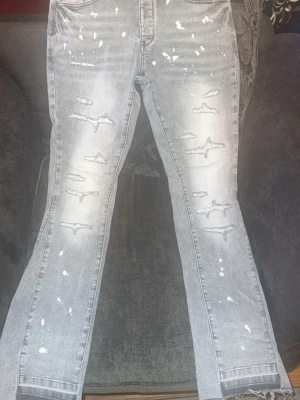 Grå Lorenzo World Flared  - Säljer ett par grå jeans frän märket Lorenzo World i modellen Oasis 2.0 med slitningar och vita färgstänk för en trendig, avslappnad look. Jeansen har raka ben, fem fickor och råa kanter vid bensluten. Perfekta för dig som gillar streetstyle och vill sticka ut lite extra. Tveka inte på höra av er vid intresse. 