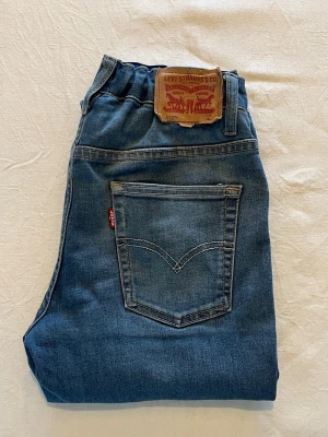 Levi's 512 blå jeans med resår - Snygga blå jeans från Levi's modell 512 med resår i midjan och klassiska bakfickor. Jeansen har raka ben och är tillverkade i denim med tydliga sömmar och läderpatch bak. Perfekta för en avslappnad och trendig look. Skadad läderpatch bak.